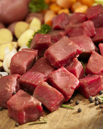 Chunks of raw stew meat on a wooden cutting board next to potatoes and vegetables