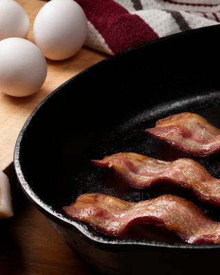 Slices of uncooked bacon and whole eggs next to a cast iron skillet of cooked bacon slices