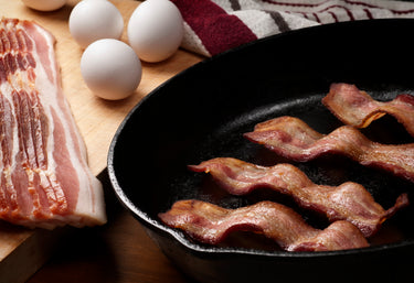 Slices of uncooked bacon and whole eggs next to a cast iron skillet of cooked bacon slices